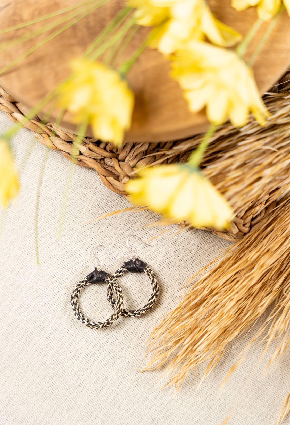 Sota Ohrringe von ABENIN – handgeflochtene Creolen aus Naturgras in Beige-Schwarz mit Lederdetail, fotografiert im natürlichen Setting mit Gräsern und gelben Blüten.